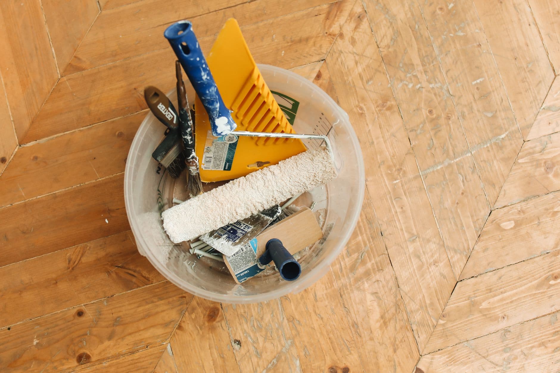 A collection of home renovation tools in a bucket on a wooden floor, ready for a painting project.